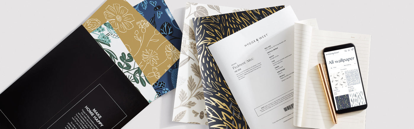 Several squares of wallpaper samples in a variety of patterns. A sheet of paper that includes Piedmont wallpaper specifications, a blank pad of paper with a copper pen and cell phone resting on top.