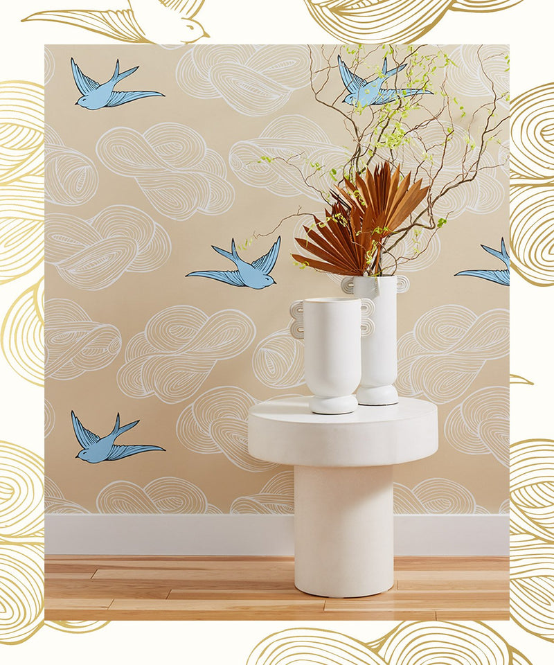 A wallpapered wall featuring a tan background with white clouds and blue birds. A white table sits in front with two white vases, one containing a dry floral arrangement. Framed around the main image is a white and gold bird and cloud pattern.